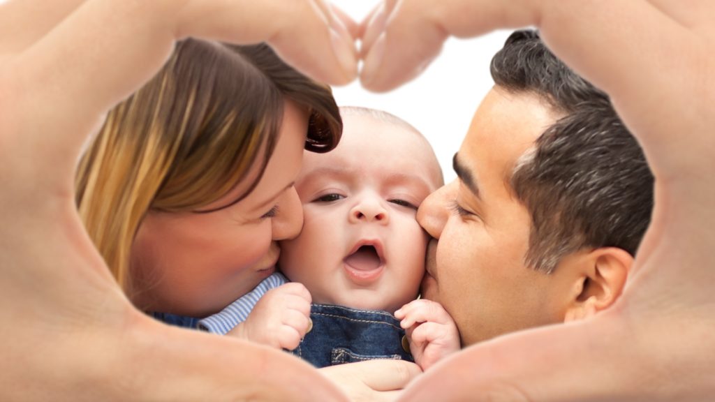 Parents kissing their baby with their hands forming a heart around them.