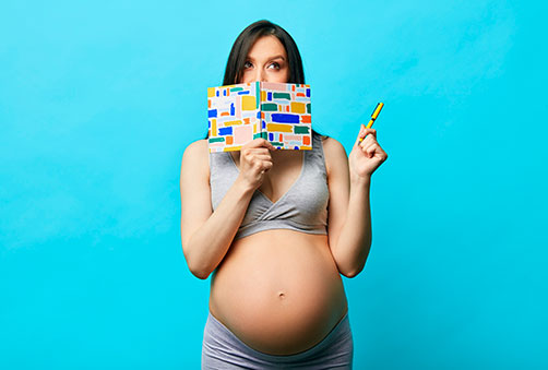 A pregnant woman covering her face with a book and pointing up.
